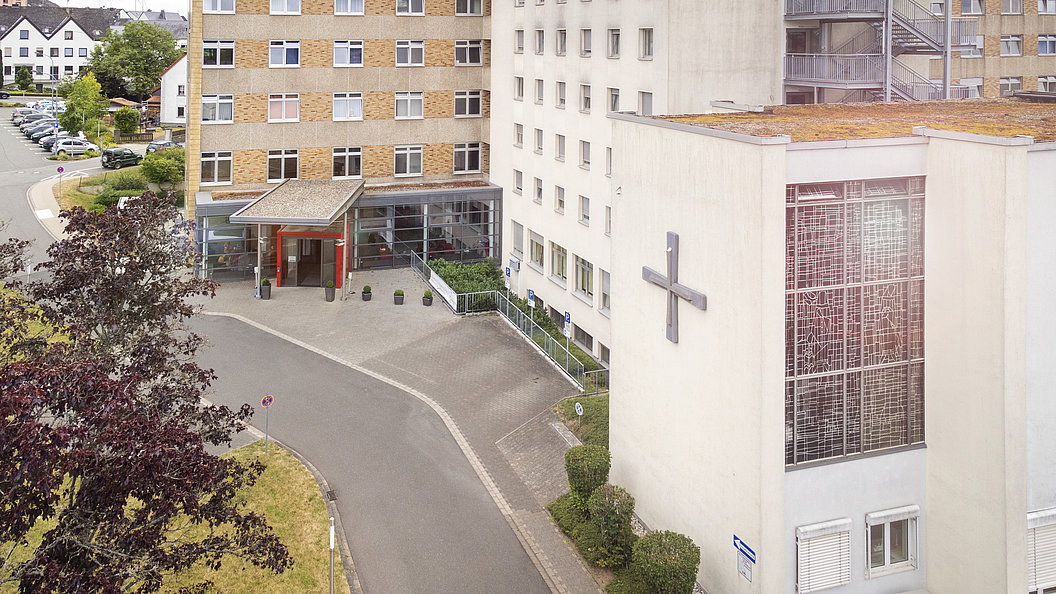 Außenansicht eines mehrstöckigen Gebäudes mit großem Kreuz an der Fassade und überdachtem Eingang, umgeben von Bäumen und Parkplätzen
