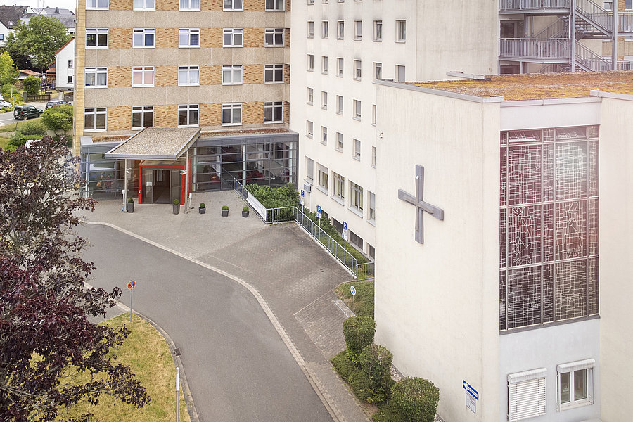 Außenansicht eines mehrstöckigen Gebäudes mit großem Kreuz an der Fassade und überdachtem Eingang, umgeben von Bäumen und Parkplätzen